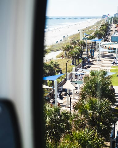 Myrtle Beach boardwalk and beach viewed from above with palm trees, shops, and ocean in the background