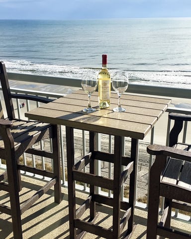 Oceanfront deck with wooden table, chairs, and white wine bottle overlooking sandy beach and calm sea