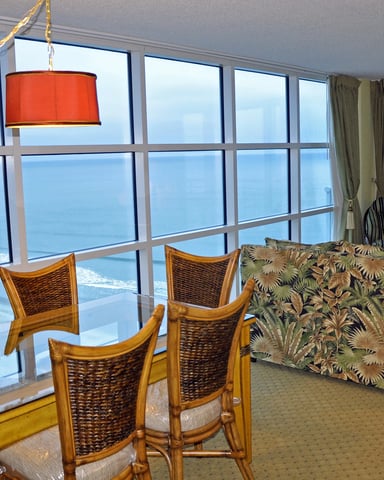 High-rise oceanfront living room with floor-to-ceiling windows, dining table, woven chairs, patterned sofa, and red pendant lamp