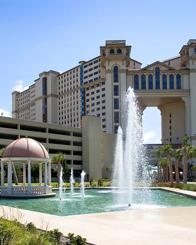 Resort courtyard with fountain, gazebo, palm trees, and large beige buildings with blue architectural details under clear sky