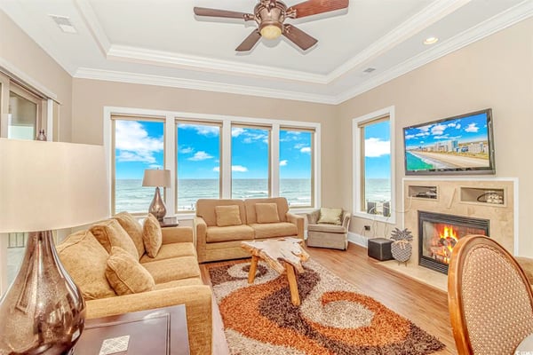 Spacious living room with gray upholstered seating, ceiling fan, blue patterned rug, and glass doors opening to balcony with ocean view.