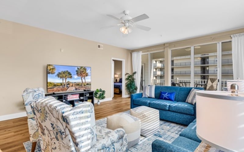 Bright living room with blue sofa, patterned armchairs, wooden floors, and glass doors opening to a balcony